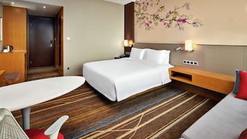 A hotel room with a large bed, a desk, a chair, a sofa, and a wall-mounted lamp.