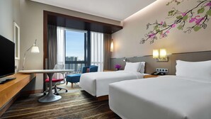 Habitación de hotel con dos camas, un escritorio con silla, televisión y vistas a la ciudad.