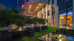 Un área al aire libre con mesas y sillas, rodeada de macetas con plantas y una entrada bien iluminada a un edificio con un letrero que dice