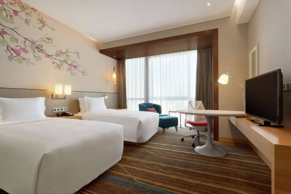 Habitación de hotel con dos camas, un escritorio con televisor, una silla y un ventanal con cortinas.