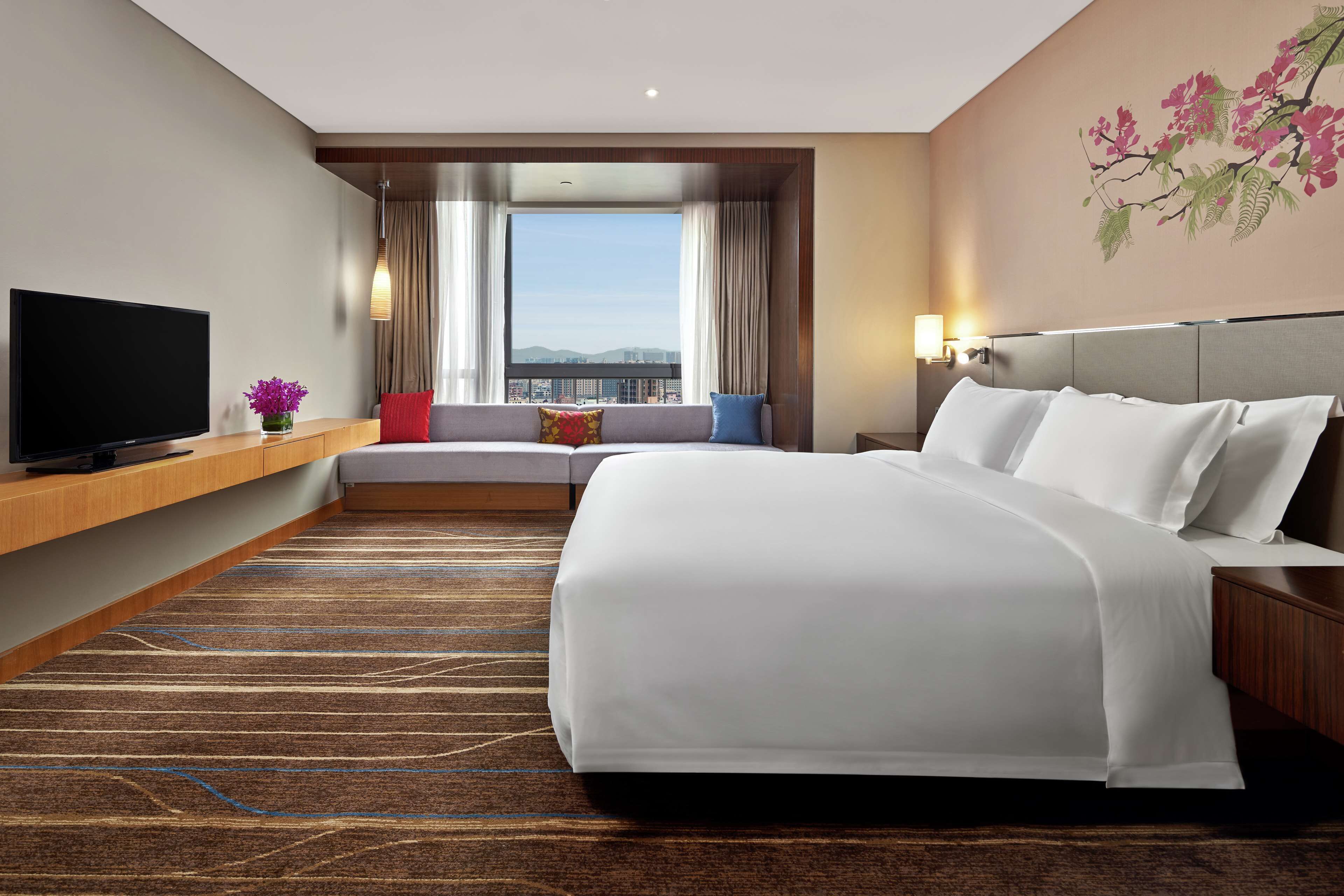 Une chambre d’hôtel dotée d’un grand lit, d’un téléviseur à écran plat, d’un canapé avec des coussins, d’une fenêtre avec des rideaux et d’un mur orné d’une fresque florale.