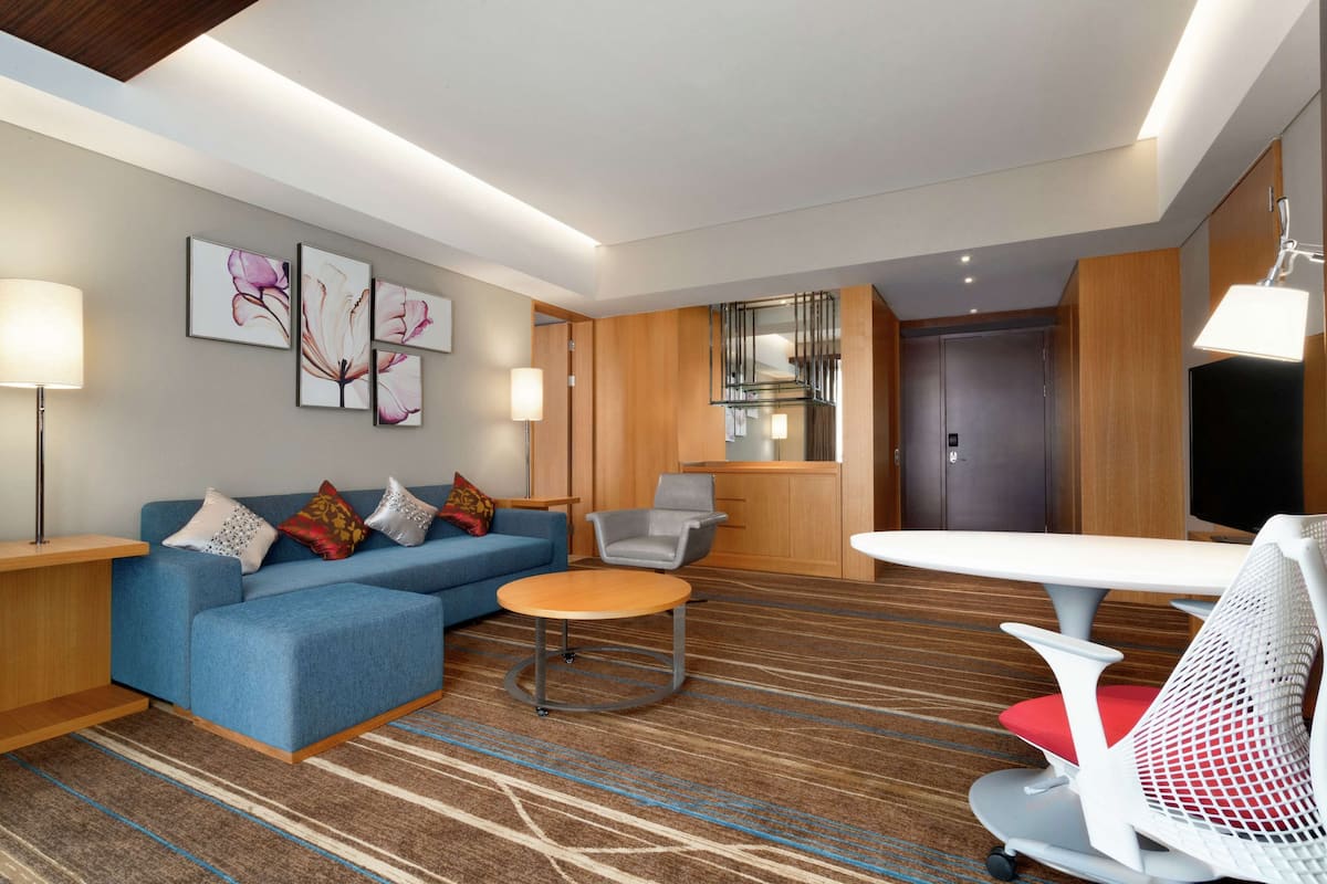Una habitación de hotel moderna con un sofá azul, una mesa de centro redonda, una silla blanca de malla y un televisor de pantalla plana.