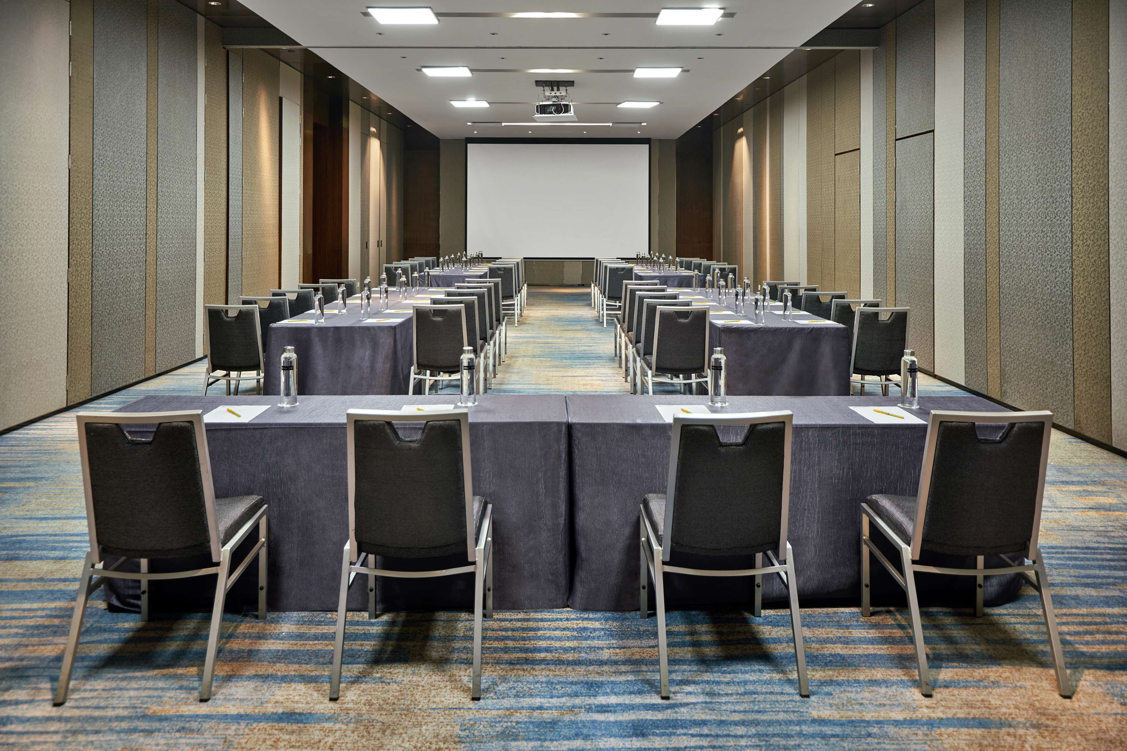 Une salle de conférence avec des rangées de tables et de chaises, un écran de projection et un sol recouvert de moquette.