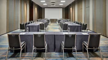 A conference room with rows of tables and chairs, a projector screen, and a carpeted floor.