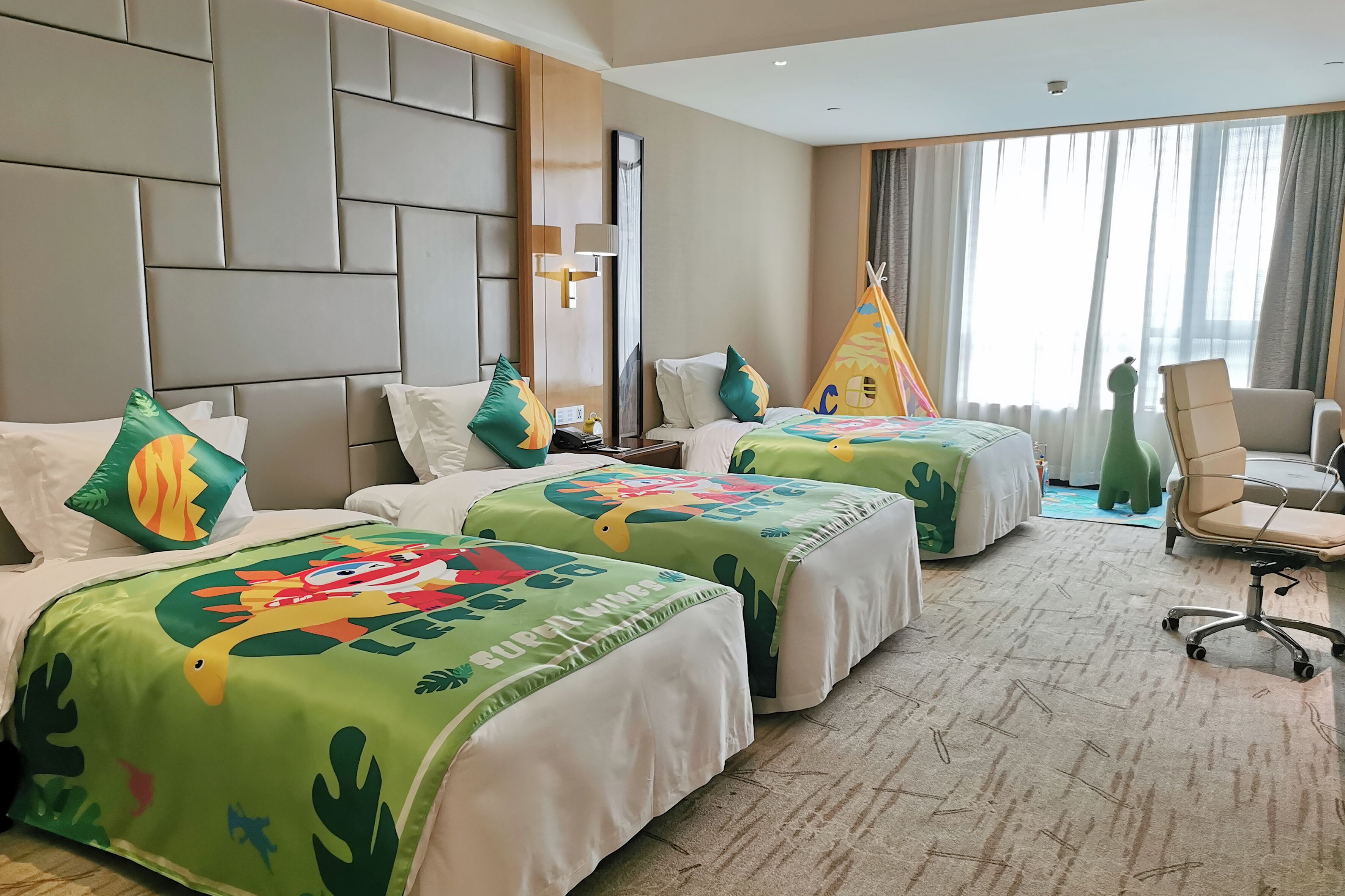 A hotel room with two beds, a large headboard, a desk, a chair, a window with curtains, and a teepee decoration.