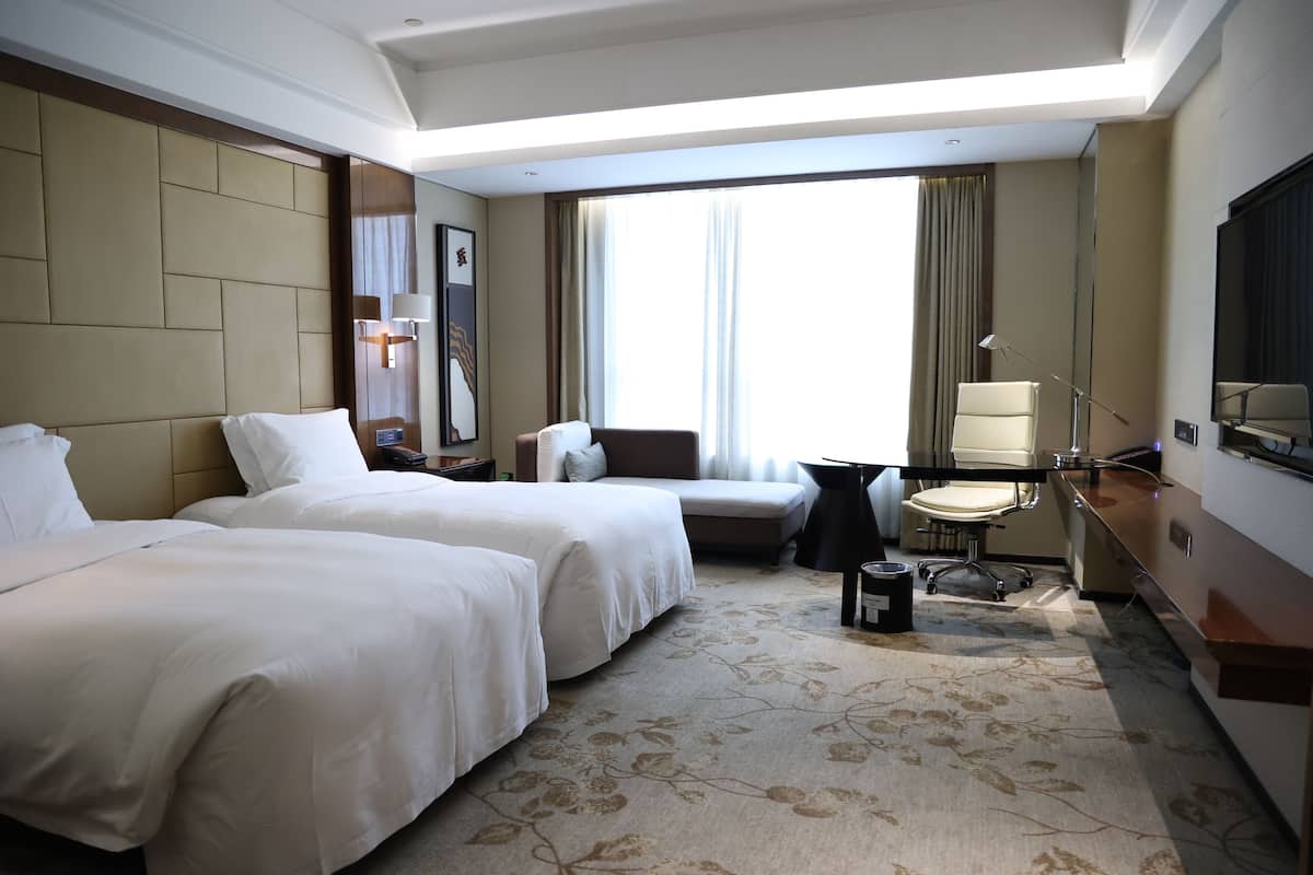 A hotel room with two beds, a desk, a chair, a TV, and a large window with curtains.