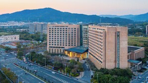 A modern hotel with a prominent glass facade, surrounded by a cityscape and hills in the background.