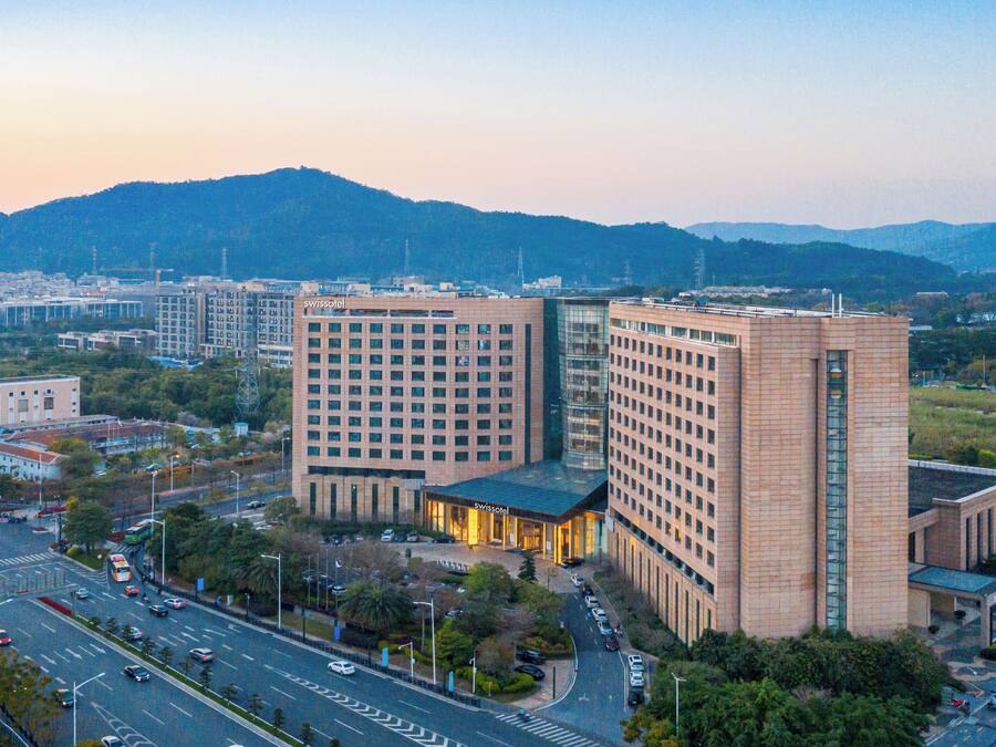A modern hotel with a prominent glass facade, surrounded by a cityscape and hills in the background.