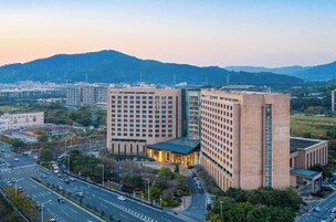 A modern hotel with a prominent glass facade, surrounded by a cityscape and hills in the background.