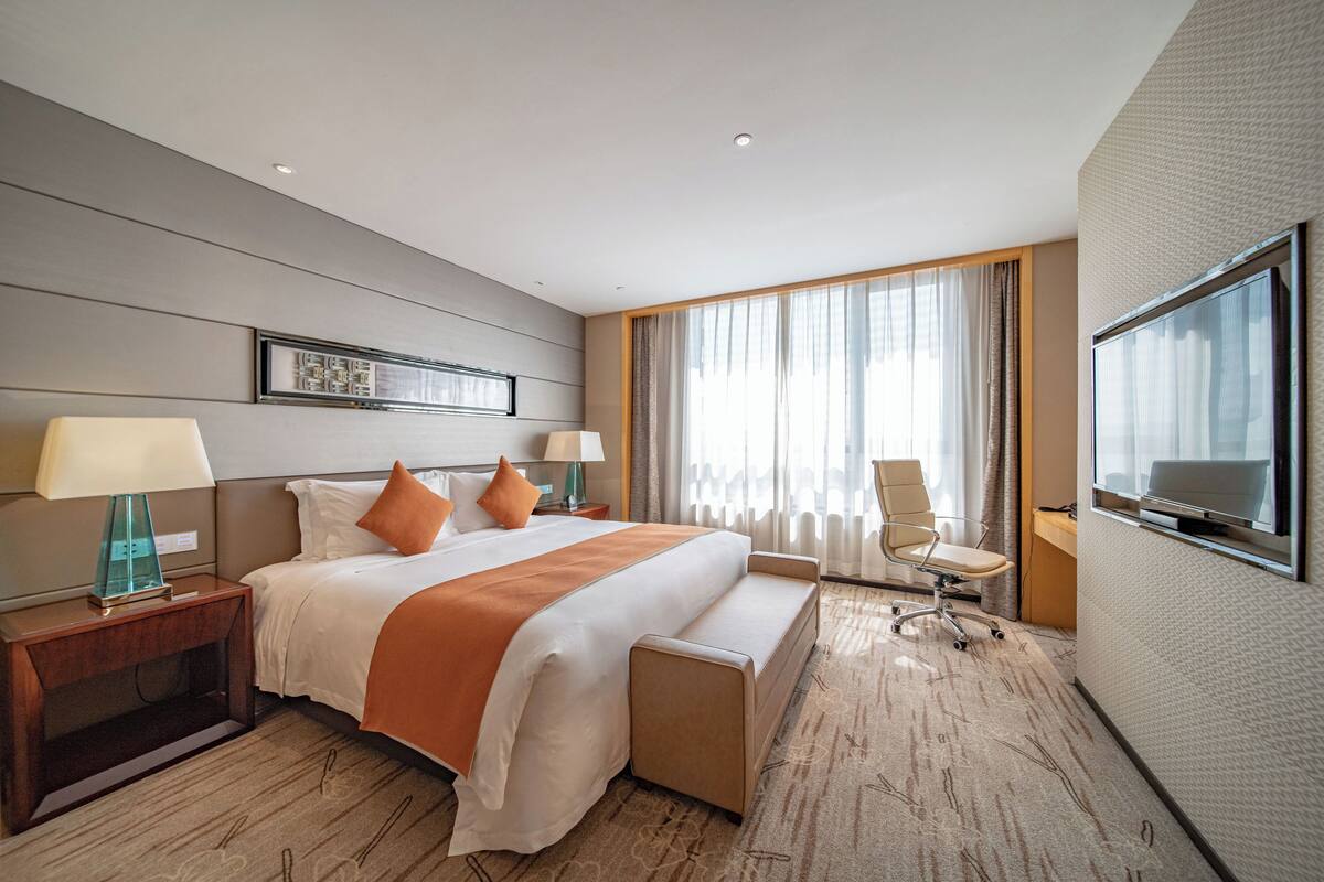 A hotel room with a large bed, two bedside tables with lamps, a desk with a chair, a large window with curtains, and a flat-screen TV mounted on the wall.
