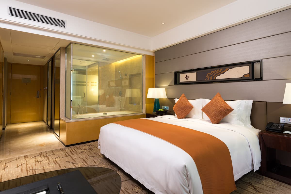 A hotel room with a large bed, a glass-enclosed shower, a bedside table with a lamp, and a framed artwork on the wall.