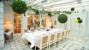 Un comedor con una mesa larga dispuesta para una comida, rodeada de sillas y decorada con plantas colgantes y faroles.