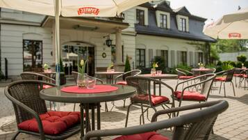 Outdoor wicker chairs with red cushions, round tables, and umbrellas at a cafe.