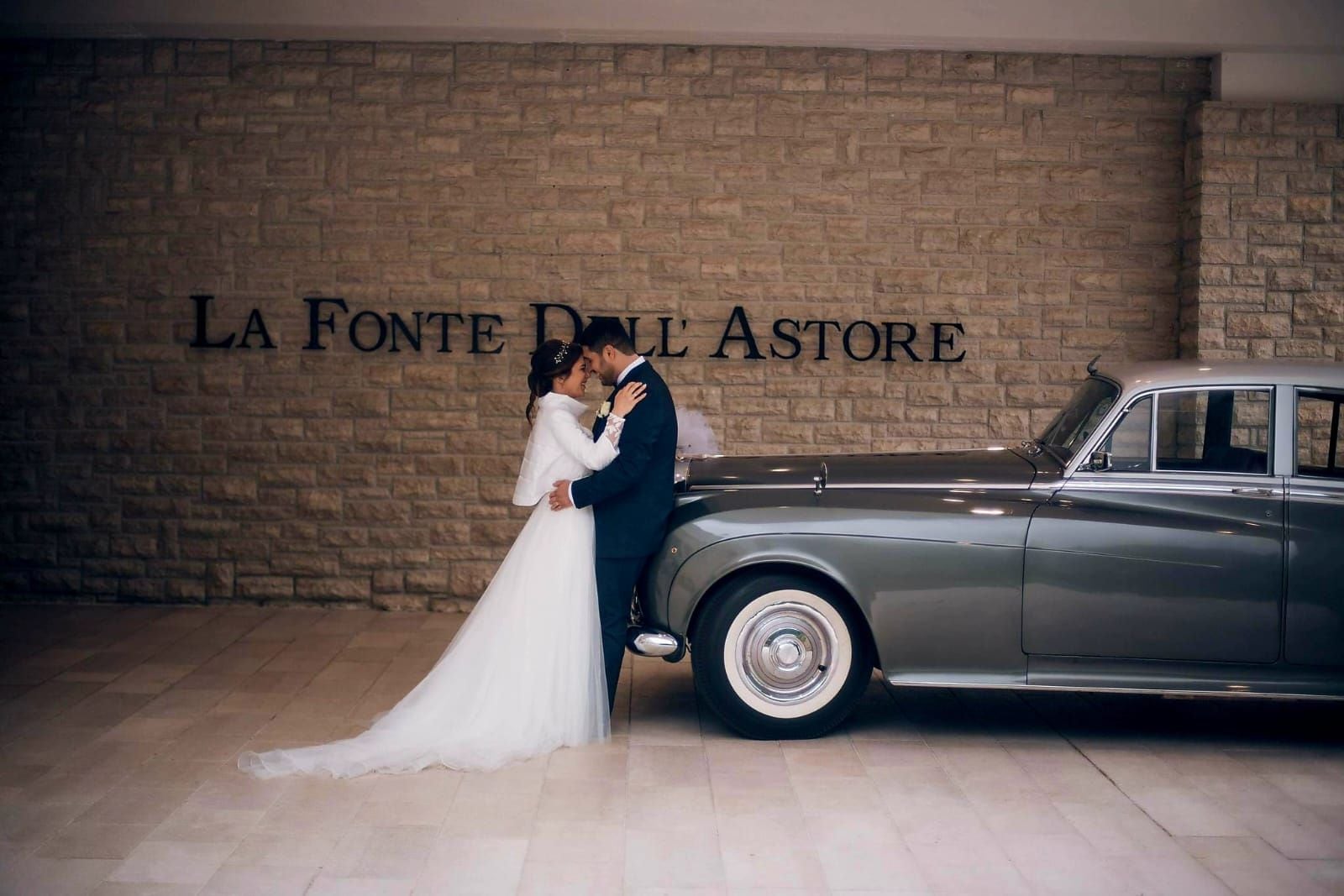 Ein Paar umarmt sich vor einem Oldtimer vor einer Ziegelwand, im Hintergrund steht die Inschrift 'LA FONTE D'ASTORE'.