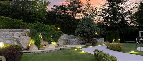 Un jardín ajardinado con muro de piedra, un sendero y diversas especies de árboles.