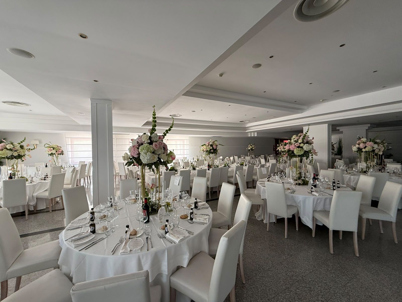 Une salle de banquet avec des tables rondes dressées pour le repas, des chaises blanches et des centres de table floraux.