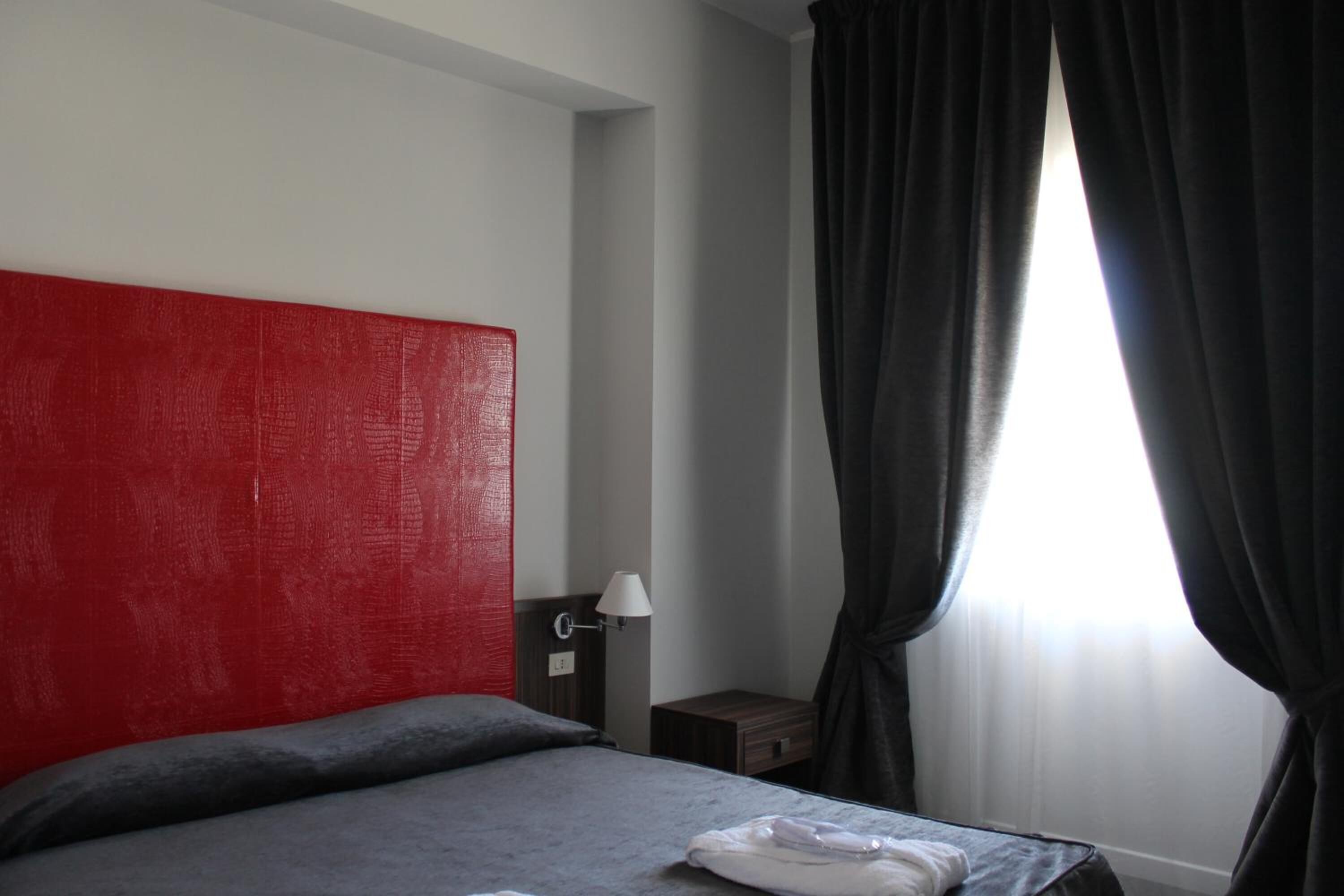 A hotel room with a red headboard, a bed with grey bedding, a nightstand with a lamp, and a window with dark curtains.