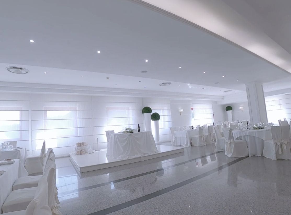 Un salón amplio con mesas y sillas blancas, ventanas grandes y un piso pulido.