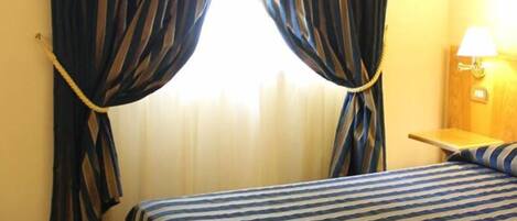 A hotel room with a bed, striped curtains, a nightstand with a lamp, and a window with curtains.
