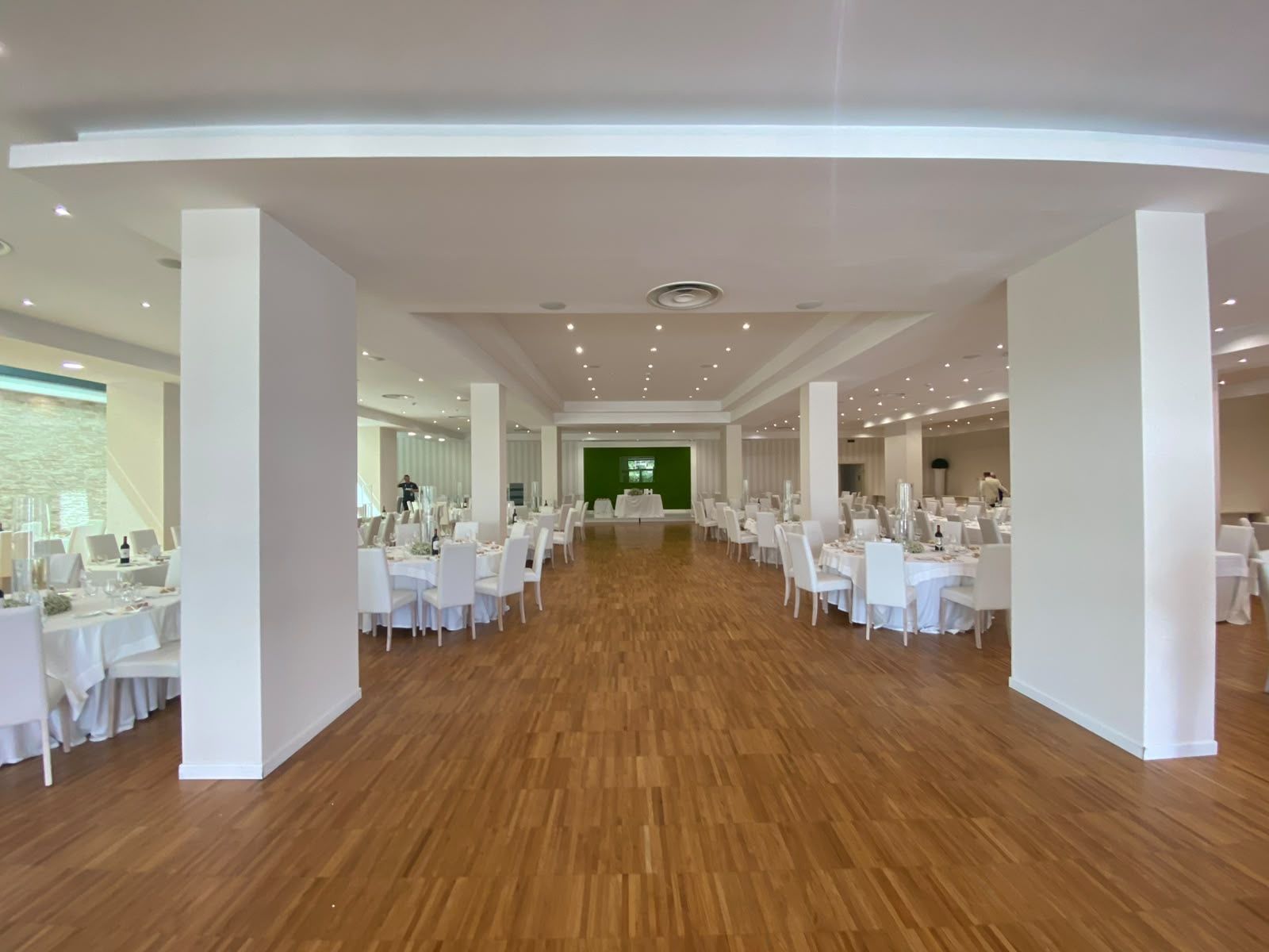 Ein geräumiger Saal mit mehreren runden Tischen, weißen Stühlen und einer grünen Leinwand im Hintergrund.