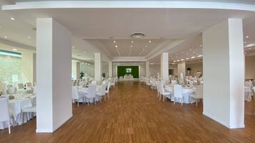 Ein geräumiger Saal mit mehreren runden Tischen, weißen Stühlen und einer grünen Leinwand im Hintergrund.