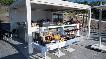 Ein gedecktes Outdoor-Buffet mit verschiedenen Speisen und Getränken, darunter Früchte, Aufgüsse und Soßen, auf einer Holzterrasse.