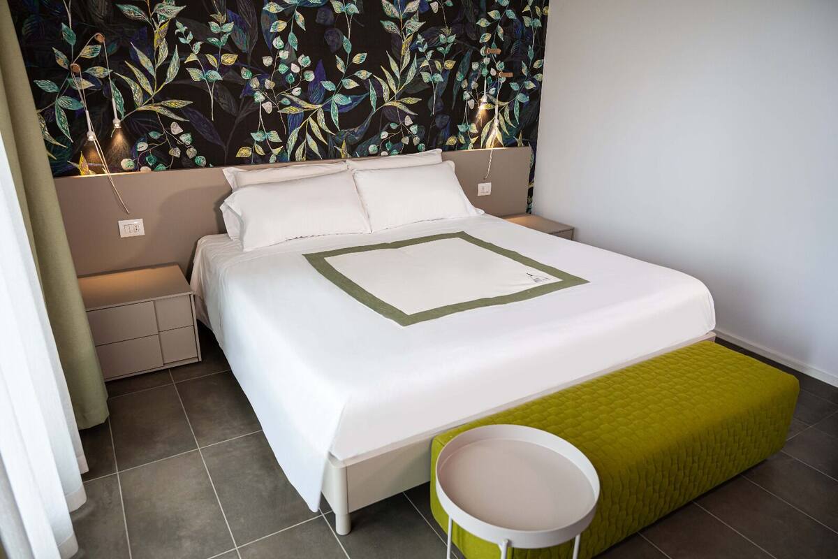 Una cama bien hecha con una colcha blanca y verde, un banco verde y una pared con estampado de hojas.
