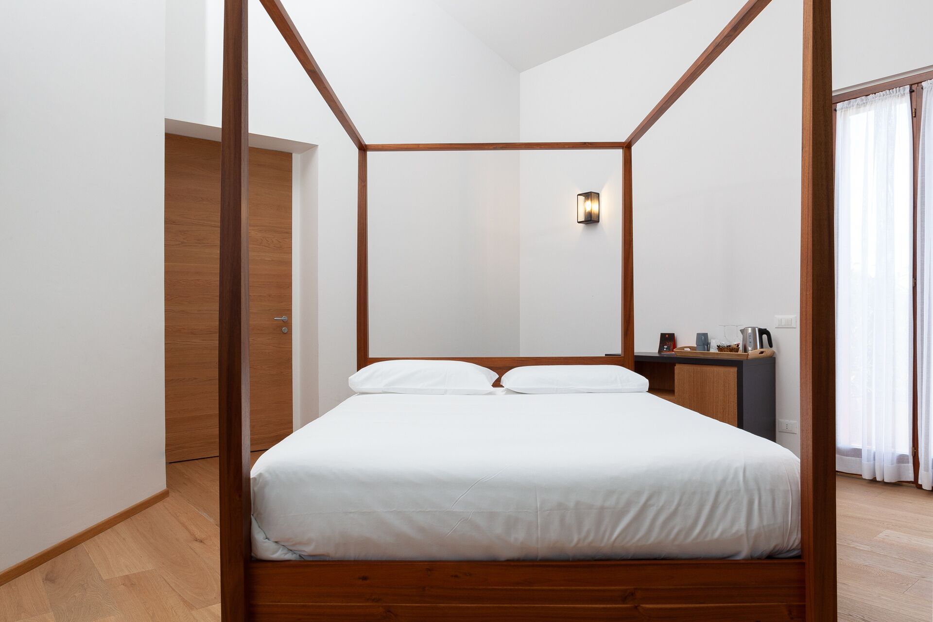 A four-poster bed with white bedding in a modern room with wooden flooring and a small bedside table.
