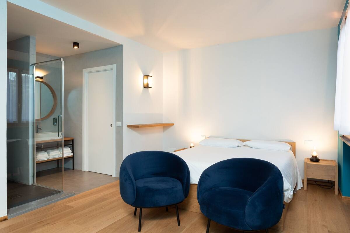 Un dormitorio con una cama, dos sillas, una mesita de noche, un velador, un espejo y una estantería.