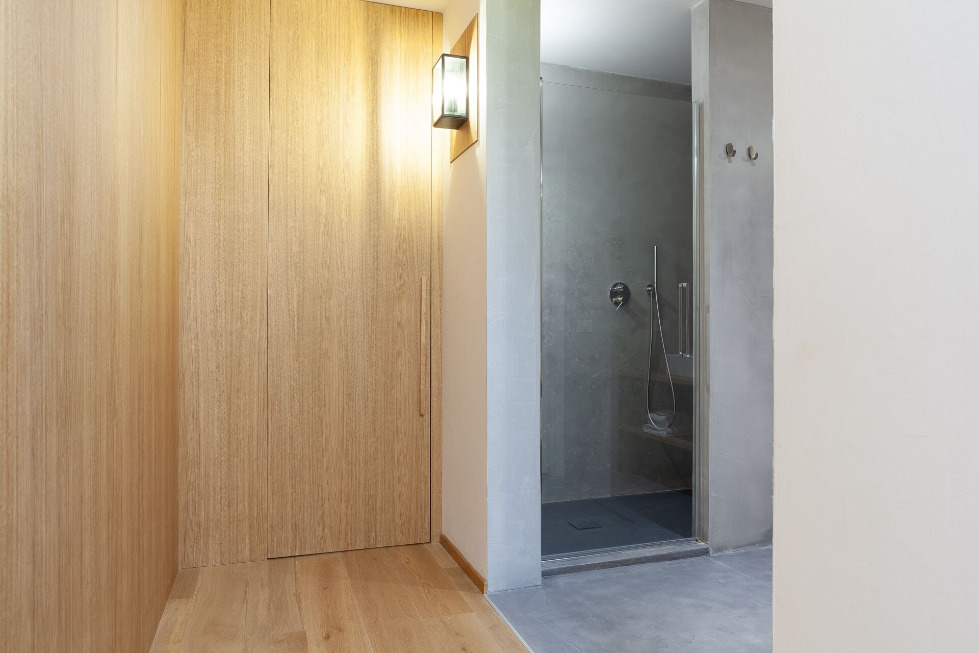 A modern bathroom with a glass shower enclosure, a wooden door, and a light fixture on the wall.