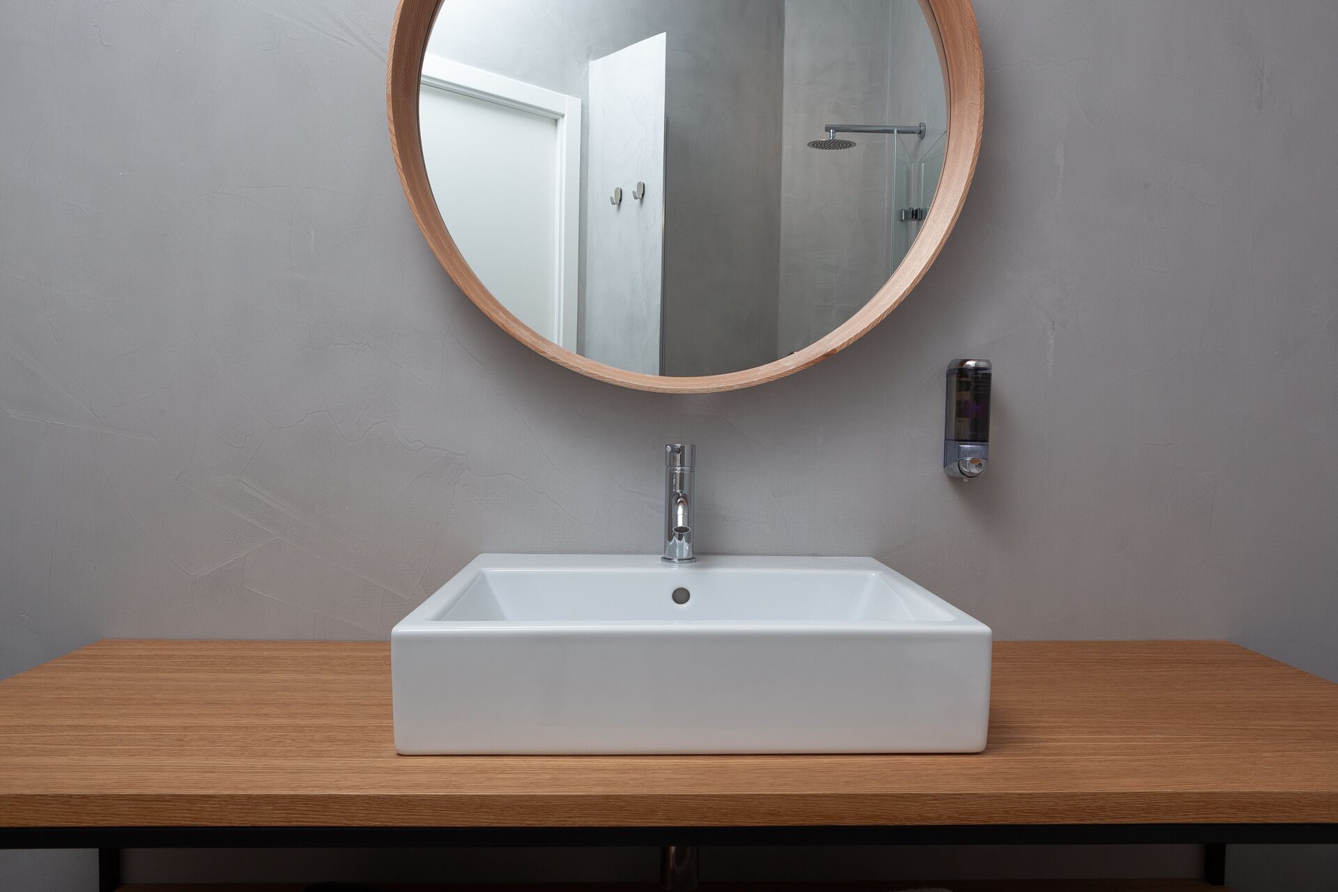 A bathroom with a rectangular white sink, a round mirror with a wooden frame, and a handheld soap dispenser on the wall.