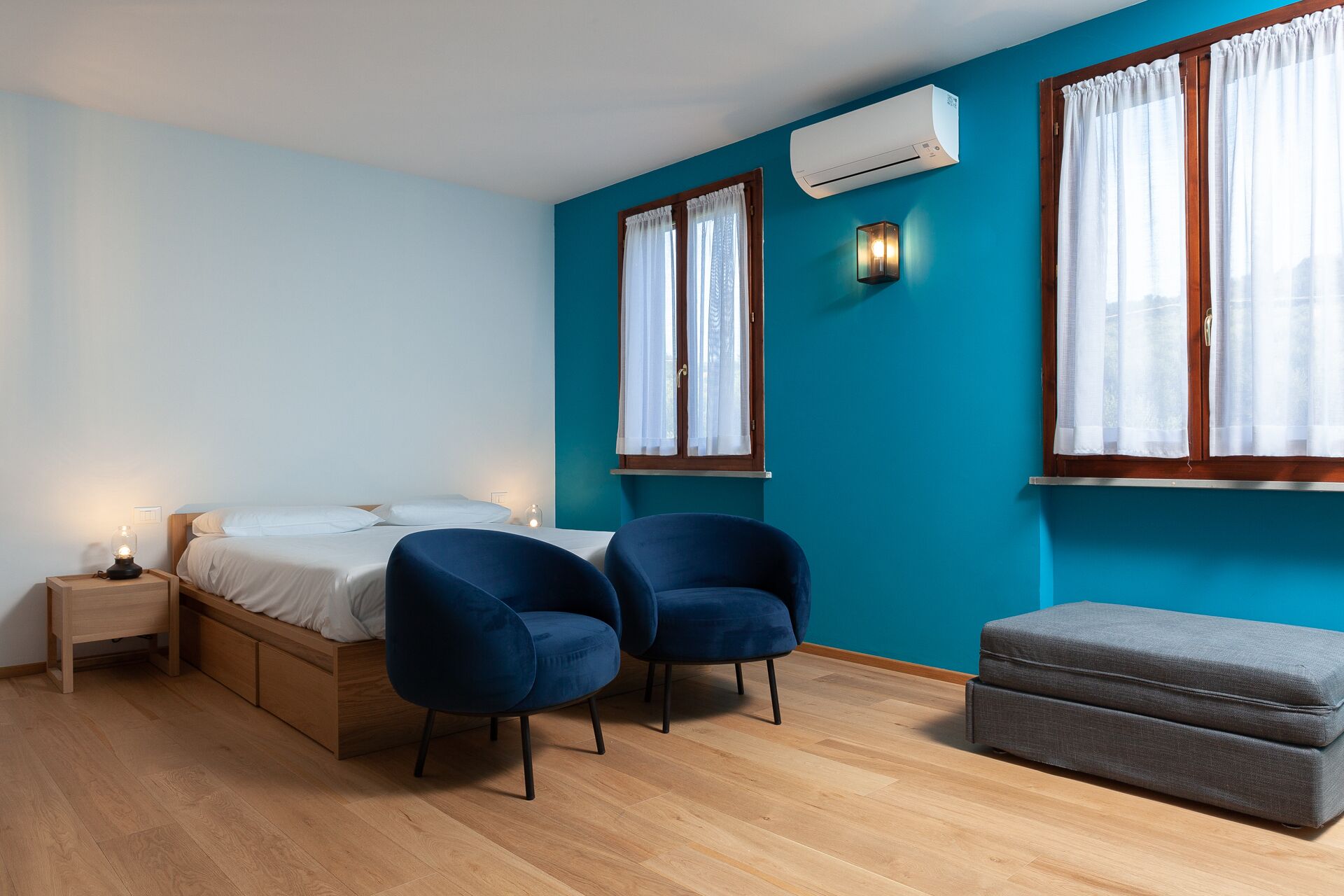 Une chambre à coucher comprenant un lit, deux chaises, un banc, un climatiseur et deux fenêtres avec des rideaux.