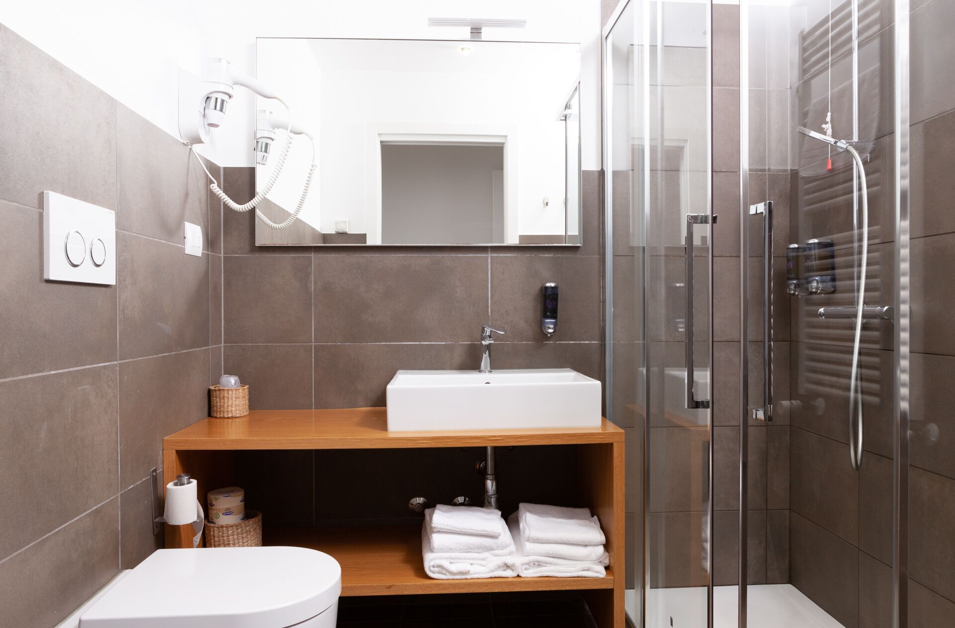 Un baño moderno con un lavabo blanco, un espejo, cabina de ducha de vidrio y un mueble de tocador de madera.