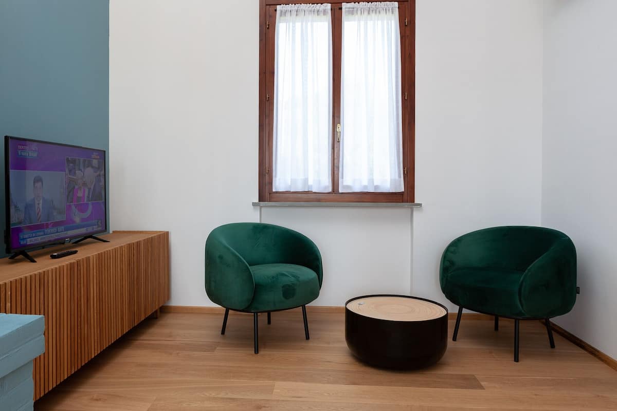 Una sala de estar moderna con un mueble de televisión de madera, dos sillones verdes, una mesa de centro redonda y una ventana con cortinas blancas.
