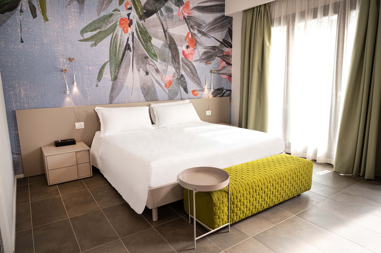 A hotel room with a bed, bedside table, lamp, and a green bench.