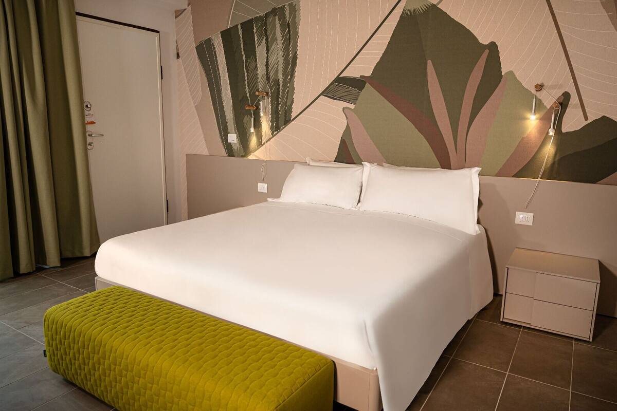 Una habitación de hotel con una cama grande, un reposapiés verde, una pared con estampado de hojas y una mesita de noche.