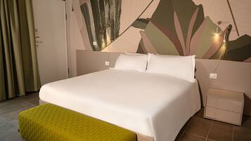 Quarto de hotel com uma cama grande, um banquinho verde, uma parede com padrão de folhagem e uma mesinha de cabeceira.