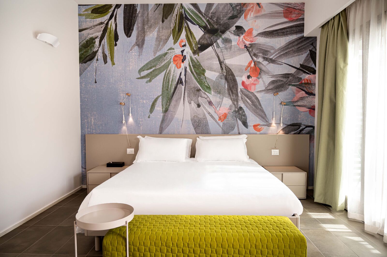 Habitación de hotel moderna con una cama grande, una mesita de noche, una pequeña mesa redonda y un llamativo mural floral en la pared.