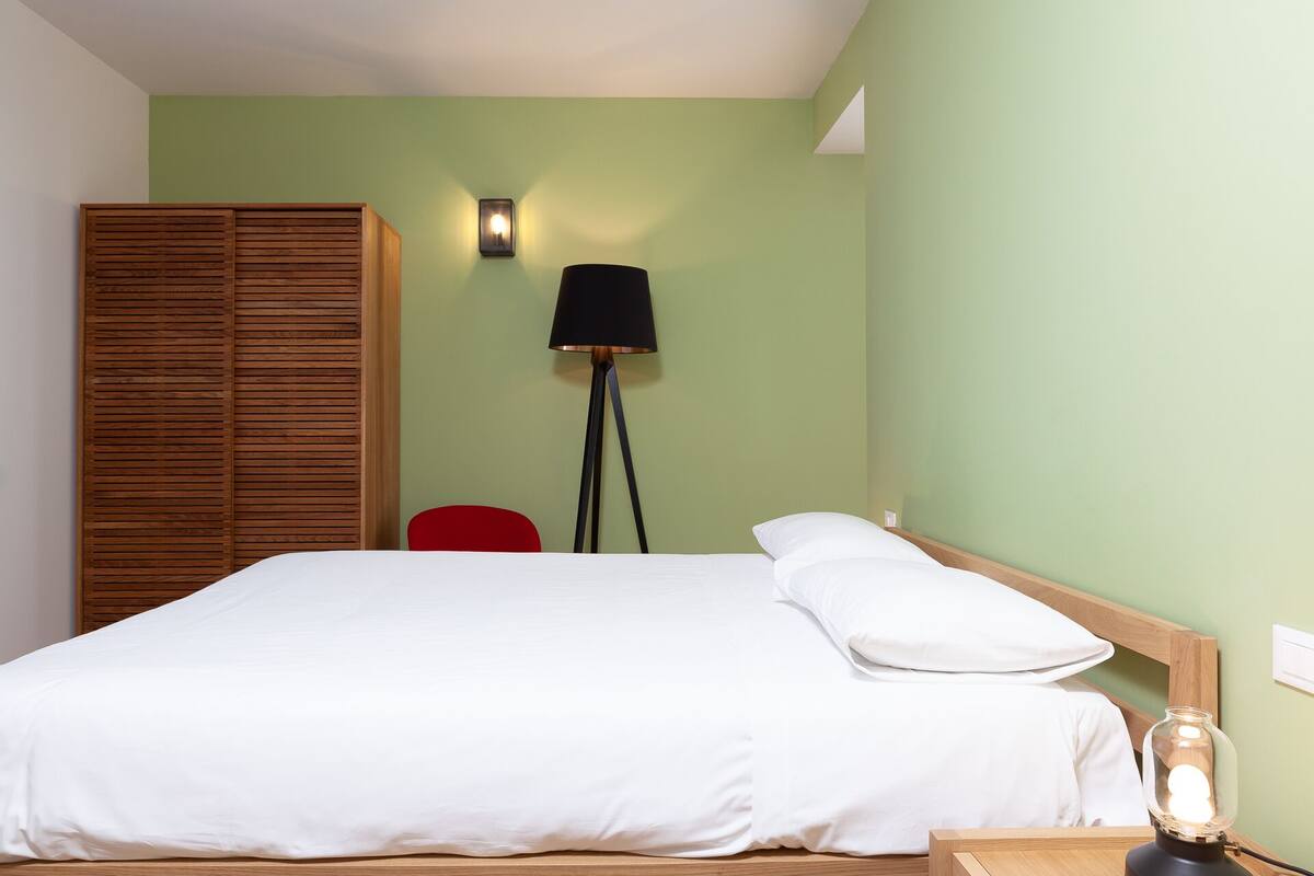 Una cama bien hecha con sábanas blancas, un cabecero de madera, una silla roja, una lámpara de pie y una mesita de noche con una lámpara.