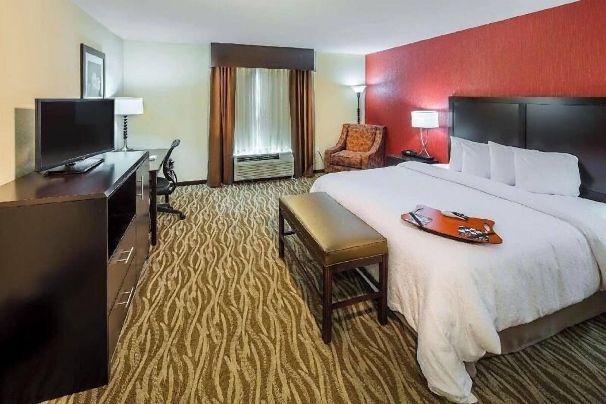 A hotel room with a bed, a desk with a television, a chair, a bench, and a window with curtains.