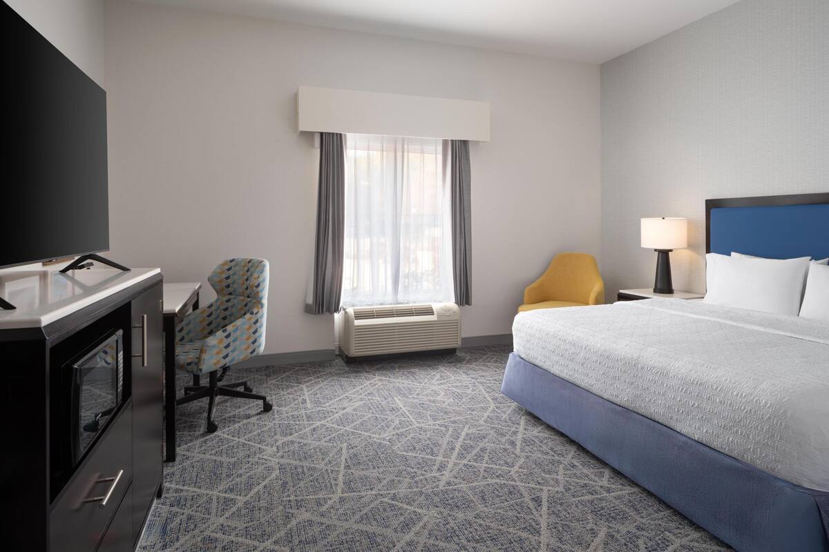 A hotel room with a bed, a desk with a chair, a TV, a lamp, and a window with curtains.