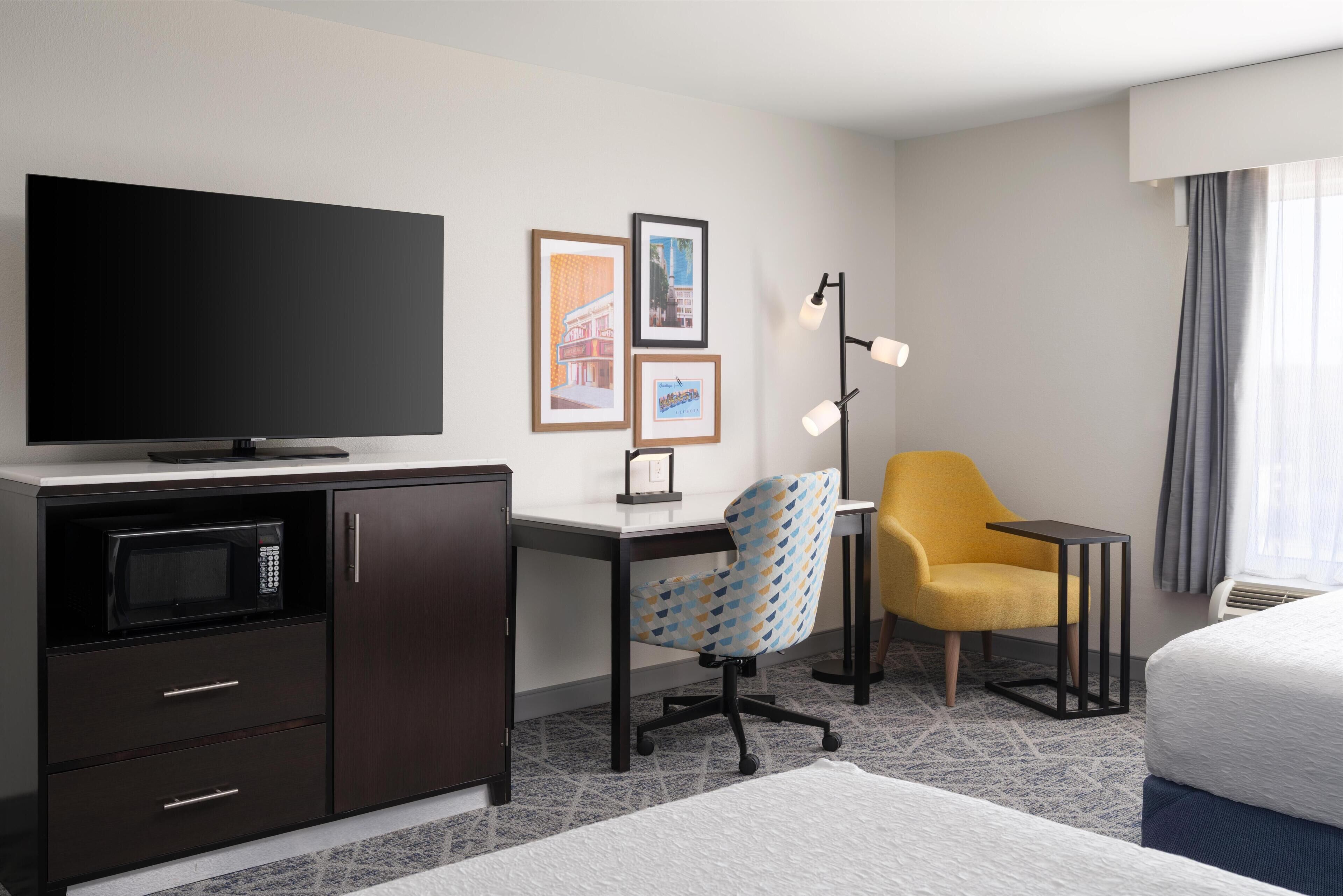 A hotel room with a flat-screen TV, a desk with a chair, a bed, and a small table.