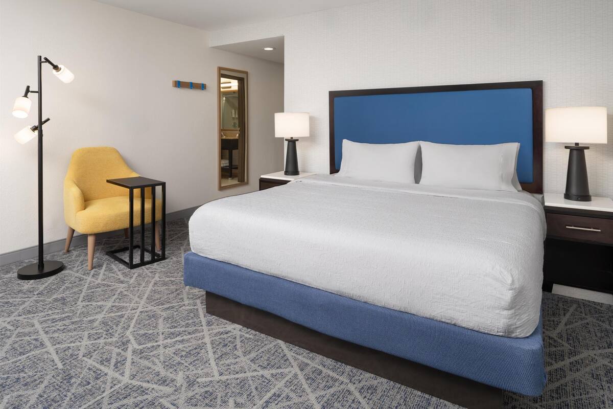 A hotel room with a large bed, two bedside tables with lamps, a small table, a yellow armchair, and a patterned carpet.