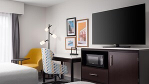 Habitación de hotel con cama, escritorio con televisor de pantalla plana, microondas, silla y cuadros en la pared.