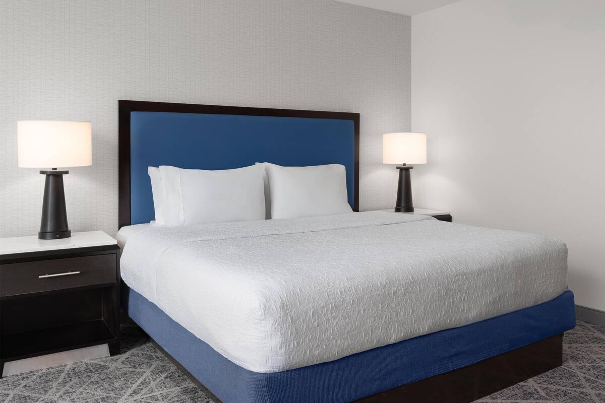 A neatly made bed with a blue headboard, white pillows, and a white bedspread. There are two bedside tables with lamps, and a dark-colored nightstand with a drawer.