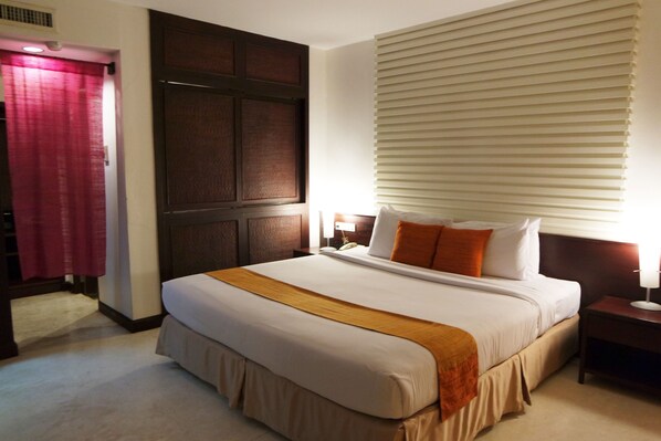 A hotel room with a large bed, a wooden headboard, a bedside table with a lamp, and a window with blinds. - Grand Hotel (Pattaya)