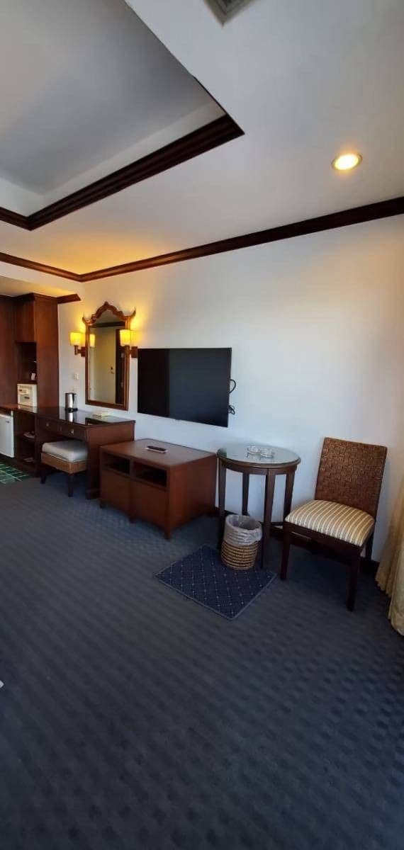 A hotel room with a flat-screen TV, a wooden desk, a chair, a small table, and a trash bin.