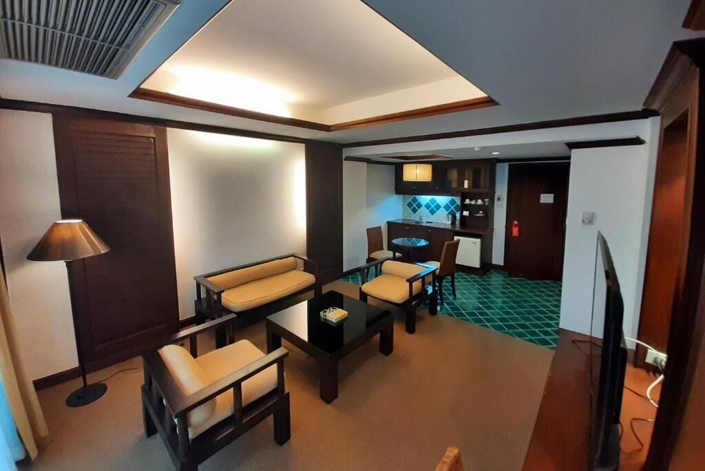 A modern hotel room with a living area featuring a sofa, coffee table, and chairs. A dining table is also visible. The room has a ceiling vent and a television.