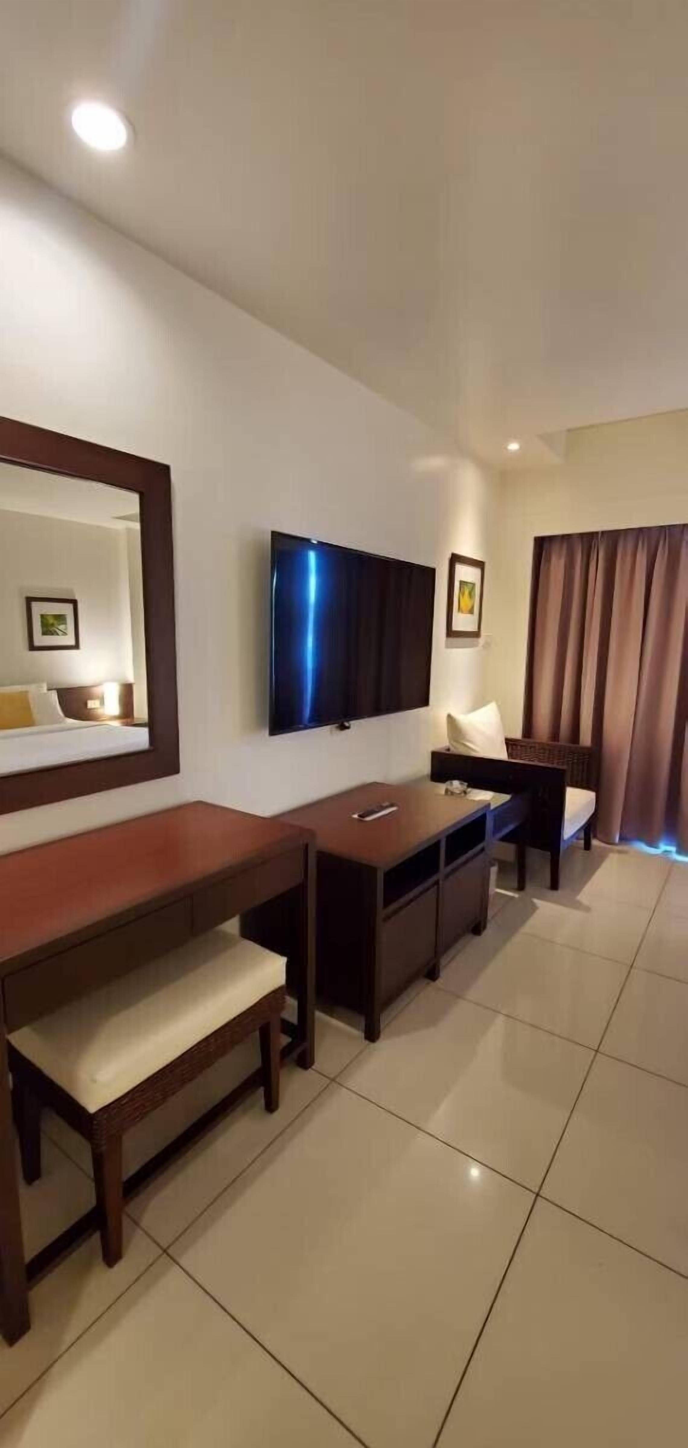 A modern hotel room with a large flat-screen TV mounted on the wall, a wooden desk with a chair, a bench, and a bed with a headboard.