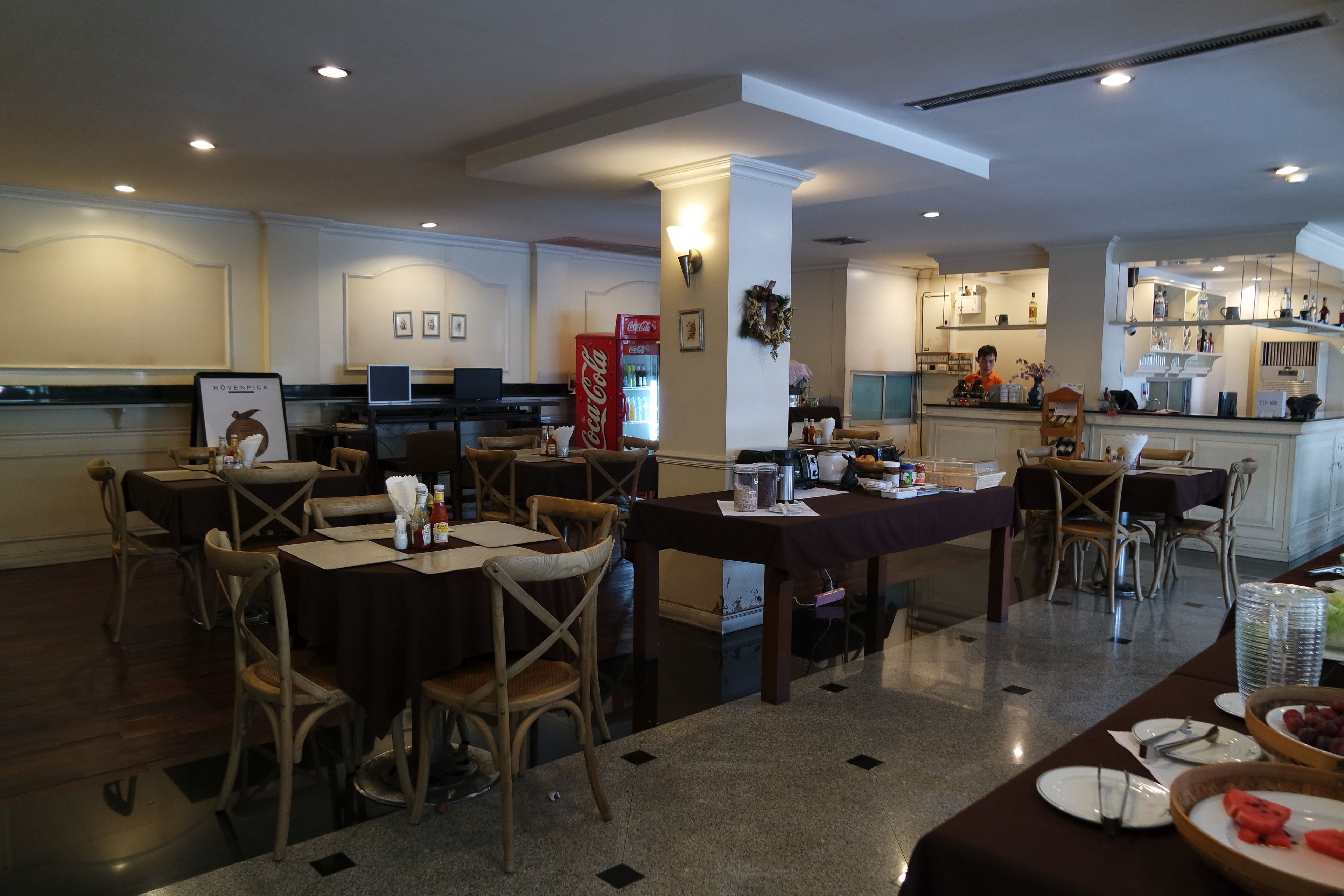 Et hotels restaurantområde med borde, stole, et Coca-Cola køleskab og et køkken i baggrunden.